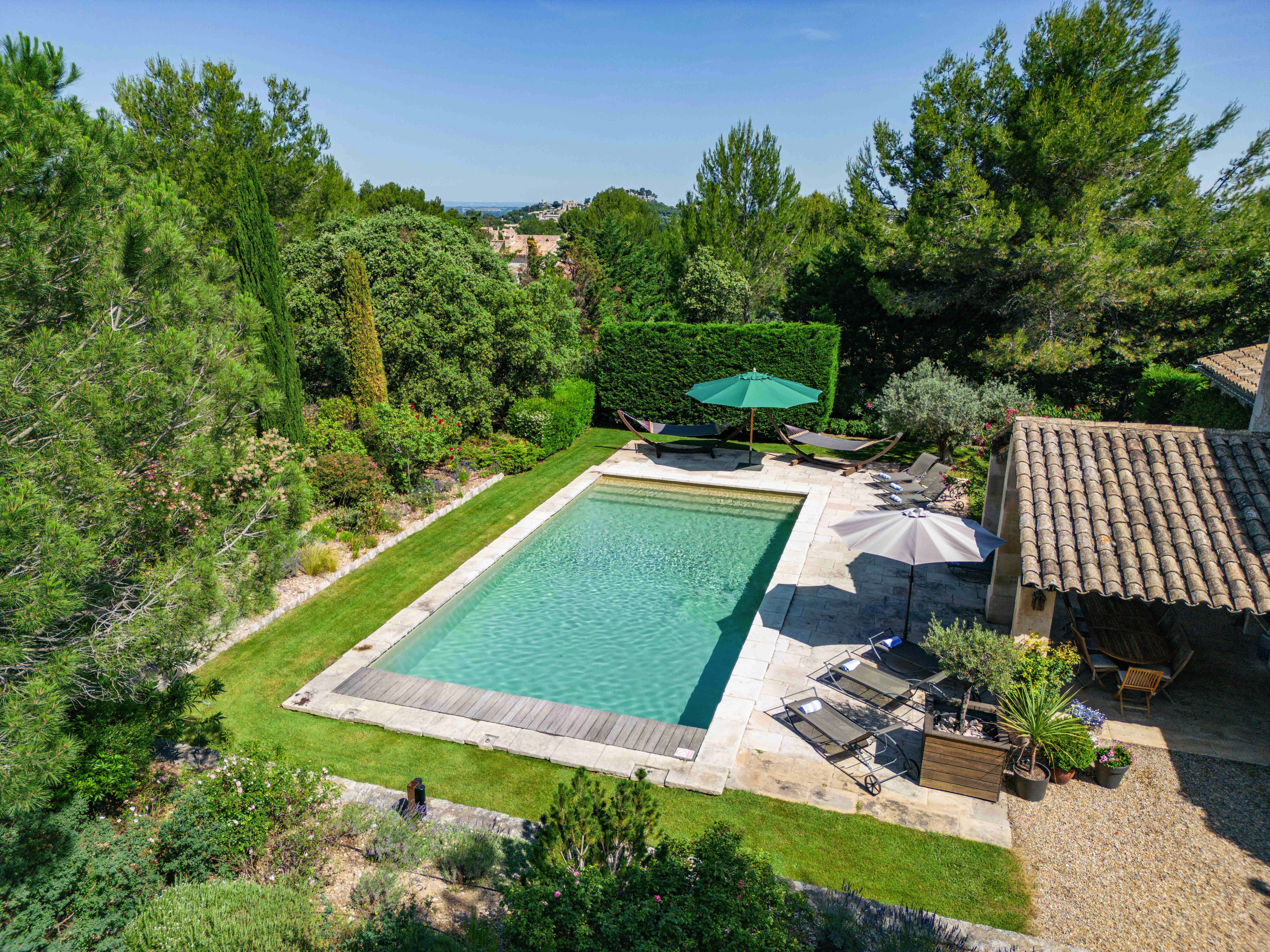 Aerial view of a serene backyard with a large rectangular swimming pool, surrounded by trees, adjacent to a stone house with a tiled roof, and outdoor furniture under green umbrellas.